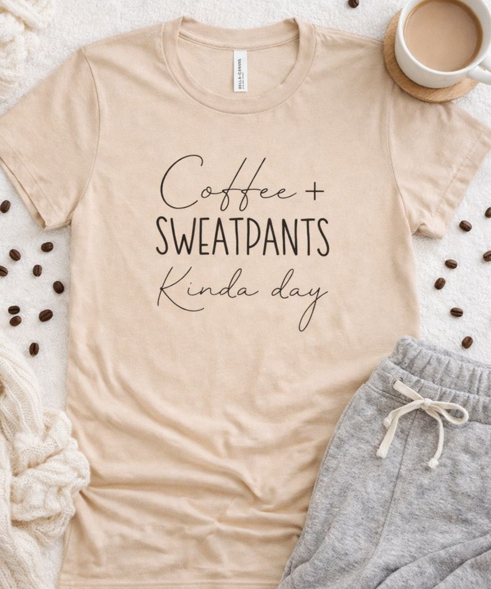 Beige t-shirt with 'Coffee + Sweatpants Kinda Day' text, paired with gray sweatpants and coffee beans on a textured surface.