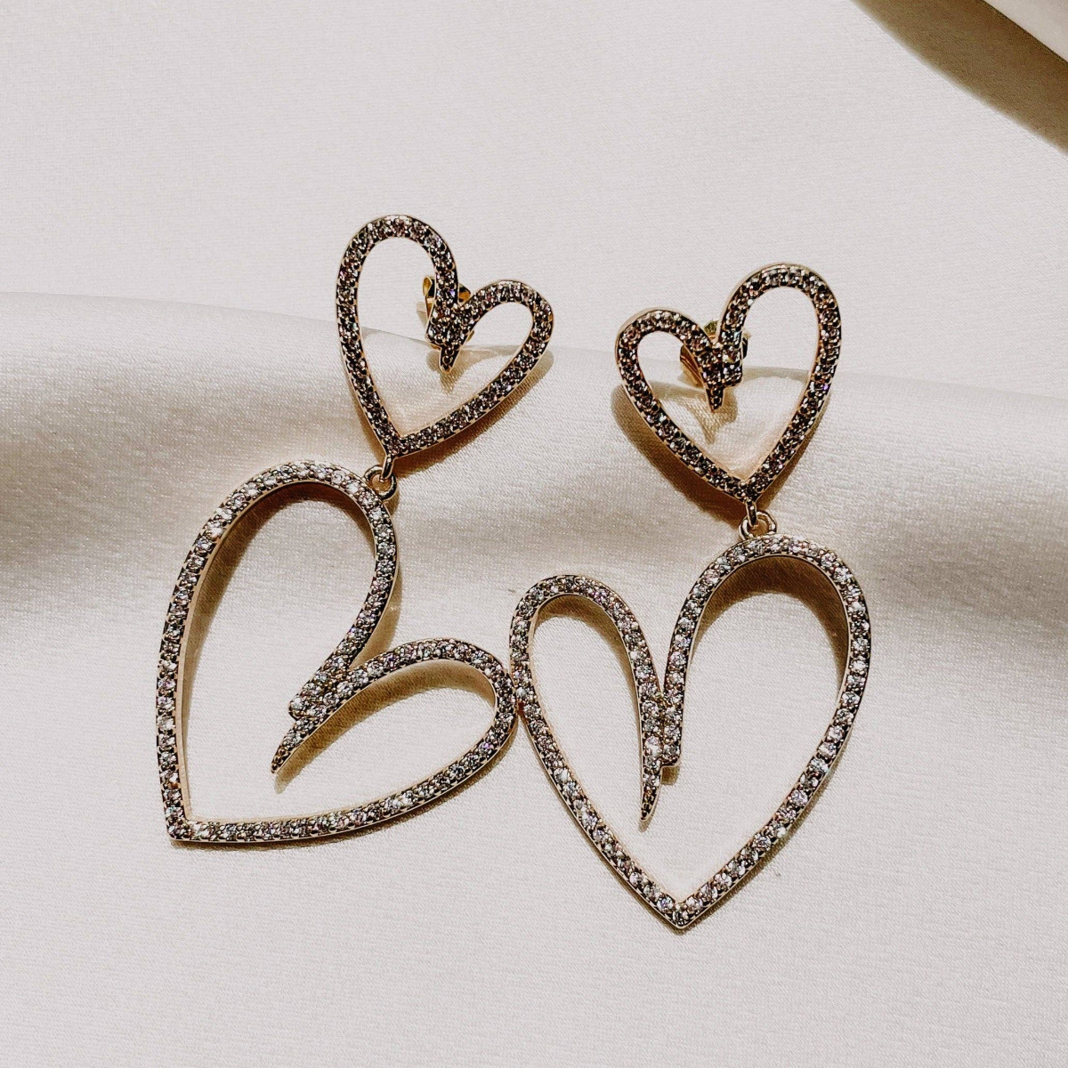 A pair of heart-shaped earrings with clear crystal embellishments on a white fabric background.