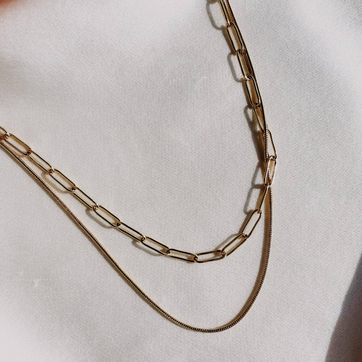A two-layer necklace with a paper clip chain and a snake chain, laid out on a white fabric surface.