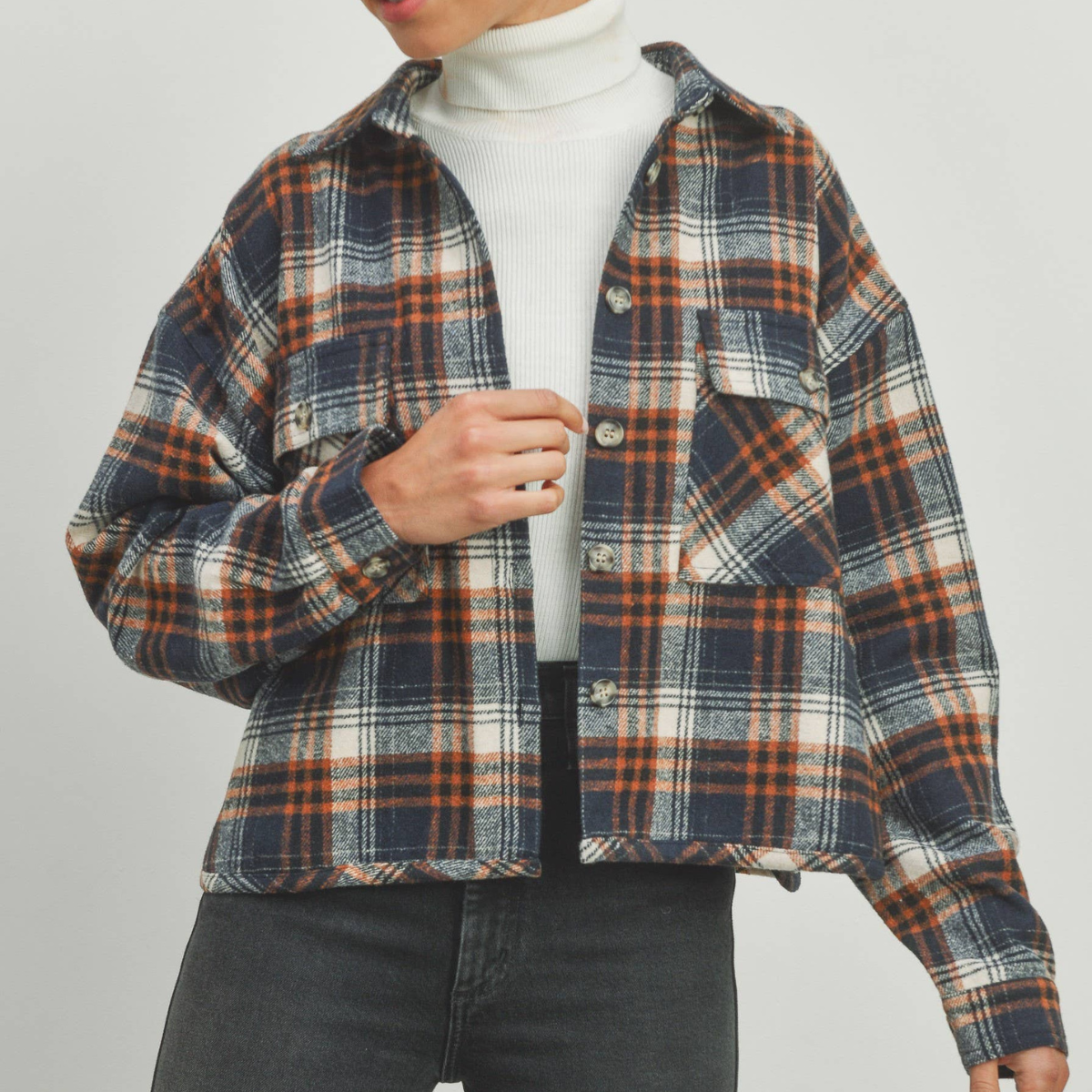 A navy blue and orange plaid cropped shacket with a classic collar and button-up front, worn by a model with a neutral facial expression and a white turtleneck underneath.