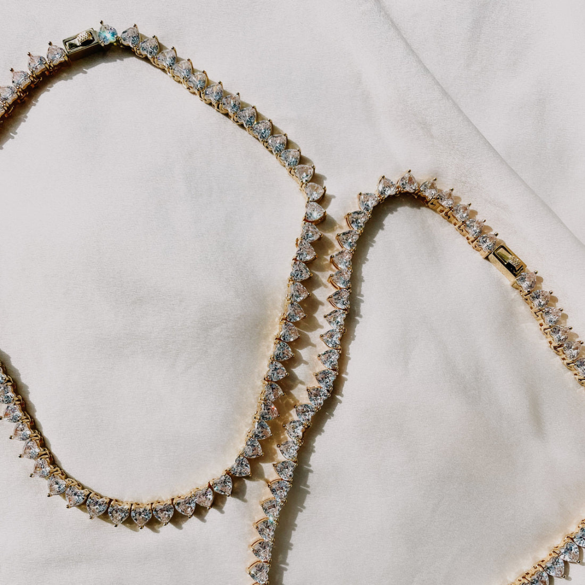 A heart-shaped necklace with blue cubic zirconia stones set in 18K yellow gold plating on a stainless steel chain, presented against a white fabric background.