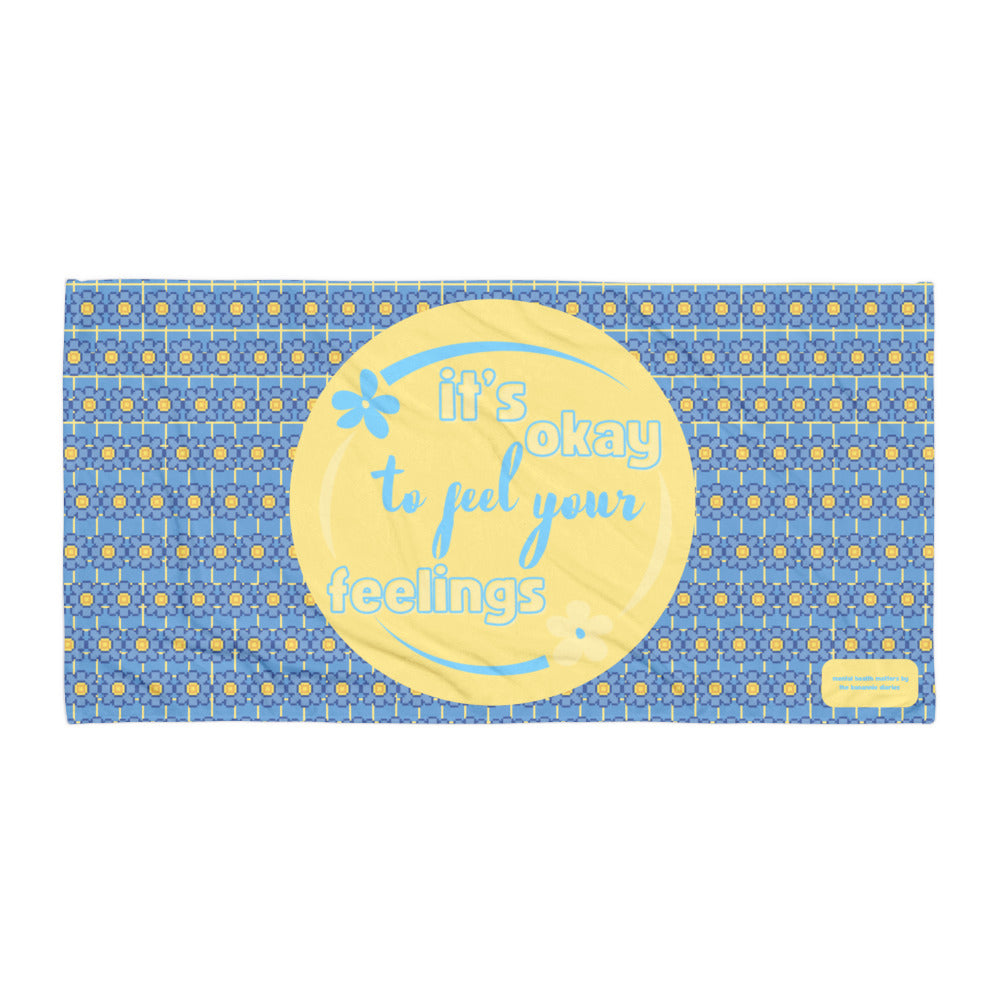 A beach/bath towel with blue and yellow flowers featuring the text 'It's Okay to Feel Your Feelings' in the center.